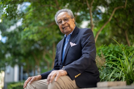 Dr Clarence B Jones in a navy blazer and glasses sits outdoors smiling, with lush greenery behind him