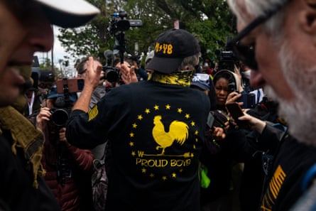 A man wearing a shirt that says ‘Proud Boys’ speaks to people holding cameras in his direction