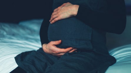 A pregnant woman sitting on a bed feeling her stomach.