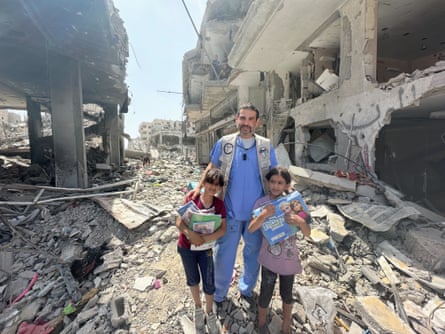 Man in blue scrubs amid complete destruction of buildings stands between two little girls holding books and a Frozen toy.