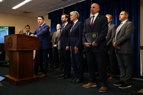 Assistant Attorney General Colin McDonald speaks at a podium while several men in suits stand in a line behind him